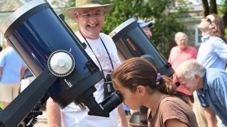 A child with brown hair in a ponytail looks into a telescope while a professor in a white t-shirt and a bucket hat looks on.