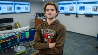 John Hill-Edgar, wearing a brown Saints Tennis sweatshirt, stands in the Bloomberg terminal with his arms crossed. There are four large televisions and two computer monitors in the background.