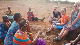 Three Saint Lawrence students sit in the desert with several local Kenyan women in vibrant clothing.
