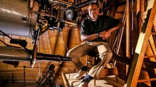 Derrik Pitts sits on a chair inside of an observatory with wood walls. A very large telescope is in front of him. 