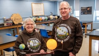 Aileen O'Donoghue and Jeffrey Miller wear brown Saint Lawrence University Gateway to the Path of Totality sweatshirts and stand in a physics lab.