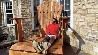A person, wearing a scarlet jacket and eclipse sunglasses, sits on a giant Adirondack chair and smiles up at the sun.
