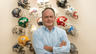 Brian McCarthy folds his arms and stands in front of a display of National Football League helmets.