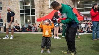 A child, wearing a bright orange sweatshirt, holds up eclipse glasses and peers at the sun while their chaperone smiles down at them. The Richard F Brush University Quad is full of students.
