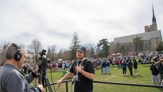 Tyler Karasinksi stands in front of a camera man as he addresses viewers on a livestream tuning into the total solar eclipse. The Richard F Brush University Quad is packed behind him.