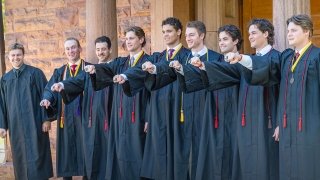 Photo shows nine men lined up with their graduation gowns on, holding out their right hand in a fist, on their hands was their Hockey championship ring.