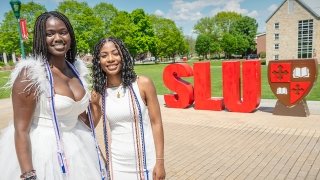 two St. Lawrence university graduating seniors pose together in front of the large Saint Lawrence University letters at the top of the quad lawn. The students have on white dresses that highlight the numerous bright colored graduation cords around their necks!