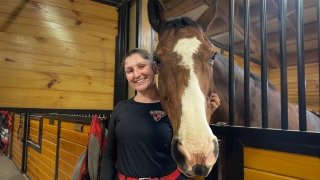 Tess Downing, wearing a black long-sleeve t-shirt, stands with her horse in the barn.