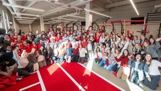 A large group of students gathers in the weight training area of Saint Lawrence University's fitness center, smiling and raising their arms in celebration. The bright, spacious gym features red and black workout equipment in the background. Sunlight streams through high windows, creating a vibrant and energetic atmosphere.