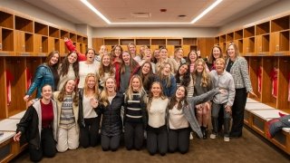 A group of Saint Lawrence University field hockey team members and staff smile and pose together in the locker room. They stand in front of wooden cubbies, with team jerseys hanging on either side. A round ceiling light displays the university’s field hockey logo above them.