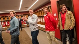 A group of Saint Lawrence University students reacts with enthusiasm and surprise while entering a locker room with rows of sports jerseys and helmets on display. They smile, clap, and look around, clearly excited by the setup. The space is brightly lit with red accents, creating a lively atmosphere.