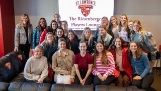 A group of Saint Lawrence University students and a staff member pose together in the Riesenberger Players Lounge, smiling in front of a screen displaying the lounge name and university logo. They are seated on leather chairs adorned with the university's crest. The setting is warm and welcoming, fostering a sense of community.