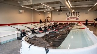 The Saint Lawrence University indoor rowing facility features multiple rowing stations with oars aligned over long water tanks for practice. A large &quot;Saints Rowing&quot; logo is displayed on the back wall. Bright overhead lights and red accents create a clean, focused training environment.