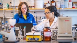 Two Saint Lawrence University students work together in a laboratory, both wearing safety glasses and focused on their tasks. One student, in a white lab coat, points to a piece of equipment while the other, in a blue lab coat, observes and takes notes on a laptop. The lab bench is filled with various scientific instruments and containers, creating a busy and engaged learning environment.