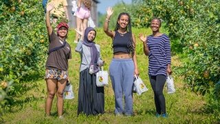 Four college students smile and wave while holding bags in an apple orchard. They are standing among green rows of apple trees, with others visible picking apples in the background.