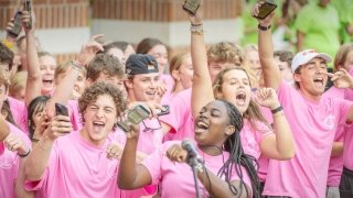 A group of Saint Lawrence University students wearing pink shirts celebrates together, many holding up their phones and cheering enthusiastically. The scene is energetic, with smiling faces and raised arms, creating a lively and joyful atmosphere. One student stands in front of a microphone, adding to the sense of excitement.