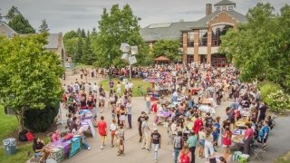A large group of Saint Lawrence University students gathers outdoors for a campus event, with tables set up displaying information and materials. Students walk between tables, engage with representatives, and socialize, creating a lively and welcoming scene. The surrounding trees and campus buildings provide a scenic backdrop.
