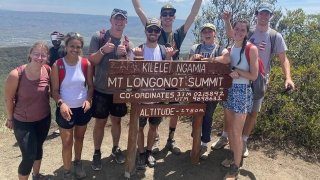 A group of Saint Lawrence University students poses around a wooden sign marking the summit of Mount Longonot in Kenya, showing their excitement and accomplishment. They smile and raise their hands in celebration, with scenic mountain views in the background. The sign indicates the altitude as 2,780 meters.