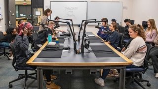 A group of students sits around a large table equipped with microphones and recording equipment in a podcasting studio. A whiteboard in the background reads &quot;Podcast Studio.&quot; The atmosphere appears collaborative and engaging, with students preparing for a recording session.