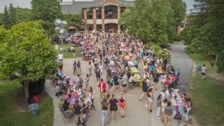 A bustling outdoor event on the Saint Lawrence University campus with rows of tables, students, and community members engaging. The scene is surrounded by trees and buildings, with colorful booths and activities along the pathways.