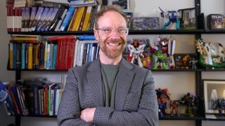 Dan Look, wearing a blazer and a green t-shirt, smiles and stands in front of shelves filled with transformer action figures.