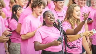 A group of students wearing matching pink t-shirts smiles and sings together, with one person holding a microphone. Many of them are holding phones, engaging with the activity. The atmosphere is lively and joyful.