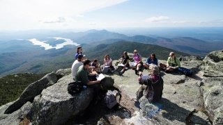 A group of students and an instructor sit on a rocky summit in the Adirondacks, engaged in discussion with notebooks and backpacks around them. The background features a sweeping view of mountains, forests, and a winding lake. The sky is clear and bright, highlighting the expansive landscape.
