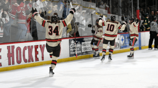 A hockey player wearing jersey number 34 celebrates on the ice with arms raised, facing the cheering crowd behind the glass. Teammates in matching uniforms skate toward the boards with raised sticks, sharing the excitement. The crowd is filled with fans wearing Saint Lawrence University apparel.