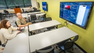 A student and an instructor sit at a desk, looking at a large screen displaying app development code and a phone interface in a computer lab classroom.