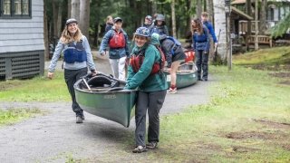 Saint Lawrence University students wearing life jackets carry canoes along a wooded path during an outdoor adventure activity.