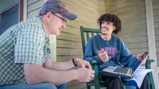 Paul Graham and Moises Peralta sit on a porch and discuss Moises's research project. Moises is holding a laptop.