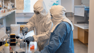 Ed Hawbeck and a professor, wearing head to toe protective jumpsuits, conduct research in a lab.