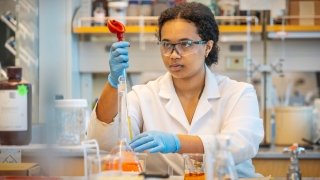 A Saint Lawrence University student, wearing a white lab coat, blue rubber gloves, and protective eye wear, measures liquid in a beaker in a chemistry lab. There are several measurement tools and beakers around them.