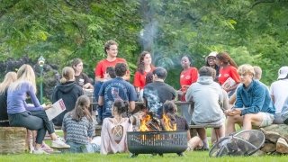 A group of students gathered around a fire pit outdoors at Saint Lawrence University, with some wearing red shirts and holding papers. They are seated on benches and logs, talking and enjoying the evening. Trees and greenery fill the background.