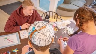 A group of kids and a young adult sit at a table covered with craft supplies and brain-themed learning materials. One child wears a paper brain cap labeled with parts of the brain. The atmosphere is hands-on and creative.
