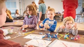 Participants color and assemble paper brain hats at a table filled with markers and supplies. Others are engaged in science activities around the room. The setting is bright and welcoming, with a focus on learning through creativity.