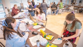 Participants color and assemble paper brain hats at a table filled with markers and supplies. Others are engaged in science activities around the room. The setting is bright and welcoming, with a focus on learning through creativity.