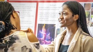 A student smiles while presenting a research poster on a travel enrichment project, engaging in conversation with a visitor during an academic showcase at Saint Lawrence University.