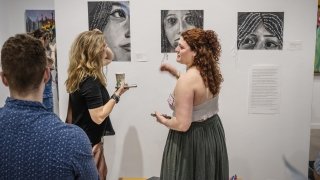 A student artist discusses her charcoal portraits with visitors at an art exhibition, featuring close-up drawings of faces with handwritten text integrated into the artwork.