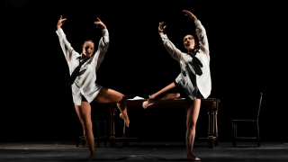 Two dancers perform on stage wearing oversized shirts and ties, striking expressive poses in front of a wooden desk under dramatic lighting