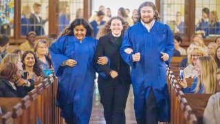 Two students in blue graduation gowns and a third in a black outfit link arms and smile as they walk down the aisle of a chapel filled with applauding guests at Saint Lawrence University.