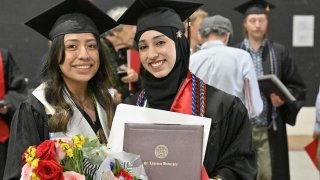 Two Saint Lawrence University graduates smile while holding their diplomas and bouquets. They are dressed in black caps and gowns, with honor cords and stoles. Other graduates and attendees are visible in the background.