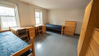 A standard double room in Gaines Hall includes two beds, two desks with chairs, and shared drawer space. Large windows bring in natural light, and the floors are covered in durable tile. The layout offers a simple and functional living space.