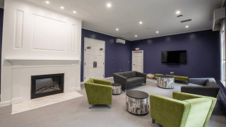 The lounge in Gaines Hall features deep blue walls, a white fireplace, and modern furniture. Green chairs, gray sofas, and patterned stools create a comfortable setting with a wall-mounted TV. Large windows on one side bring in natural light.