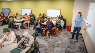 A professor leading a class from the front of the room, while students sit around tables.
