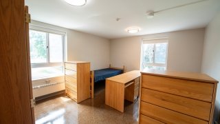 A single room in Lee Hall includes one bed, a desk with chair, and two dressers. The space has neutral walls, tile flooring, and two large windows for natural light. It offers a quiet, private living area.
