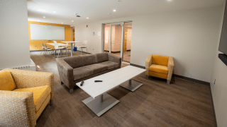 The lounge in Lee Hall includes a mix of soft seating, a white coffee table, and a wall-mounted TV. A study area with white tables and chairs sits in the back, along with a large whiteboard on a yellow accent wall. The space is open and bright with wood-style flooring throughout.