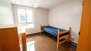 A compact single room in Lee Hall includes one bed, two dressers, and a large window for natural light. The space has neutral walls and polished tile flooring. It offers a quiet and private environment for one student.