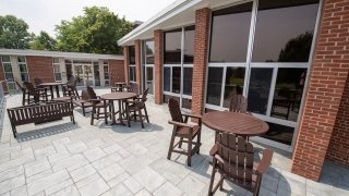 Outdoor patio at Lee Hall with round and rectangular wooden tables and chairs, next to a red-brick building with large windows.