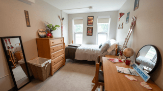 Decorated single room with a bed, wooden dresser, desk, chair, and personal items like posters, skis, and a mirror.